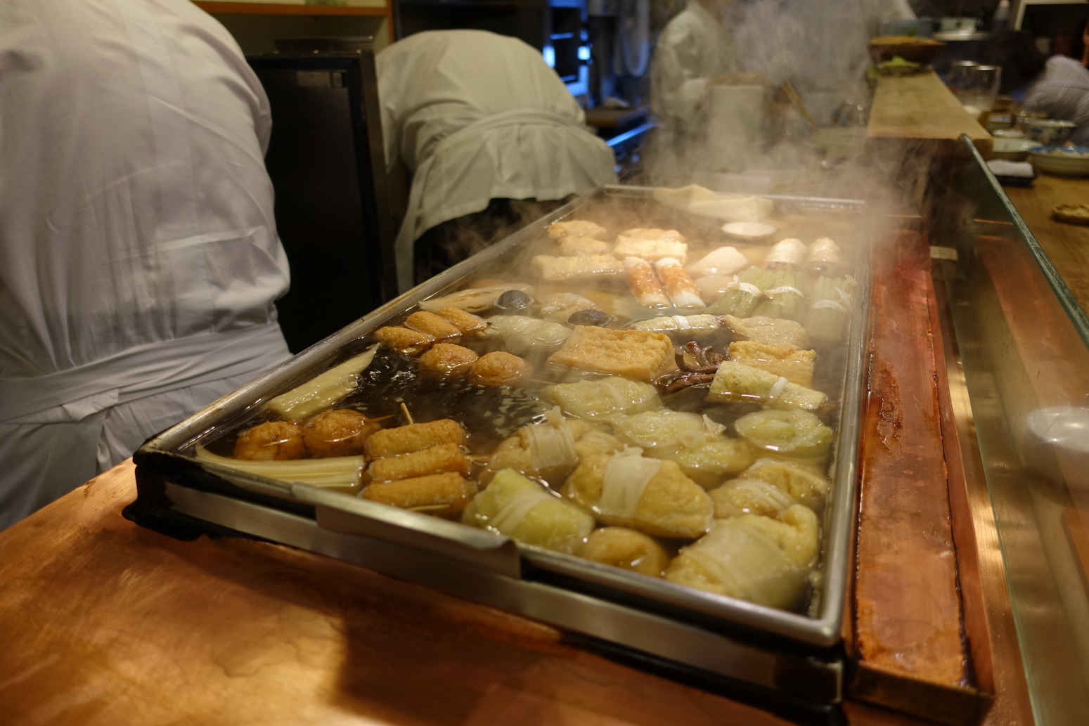 Long tray of boiling broth with parcels of vegetables and skewered meats